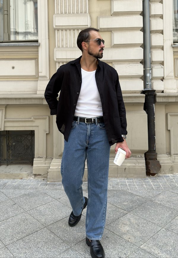 Mann mit Sonnenbrille, schwarzem Hemd, weißem T-Shirt, blauer Jeans und schwarzen Schuhen geht auf dem Bürgersteig der Stadt und hält einen weißen Kaffeebecher.