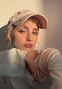 Cappellino rosa chiaro con dettagli ricamati, maglia a maniche lunghe bianca a coste, trucco naturale e capelli acconciati. Mano appoggiata al mento.