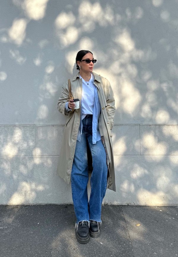 Trench coat bej peste o bluză albastră și un maieu alb, combinat cu blugi largi albaștri și încălțări cu platformă negre. Ținând o ceașcă de cafea.