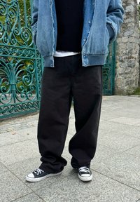 Sort løse bukser, denimjakke og sorte og hvide sneakers. Tekstur: glat stof, synlige syninger og kontrasterende hardwaredetaljer.