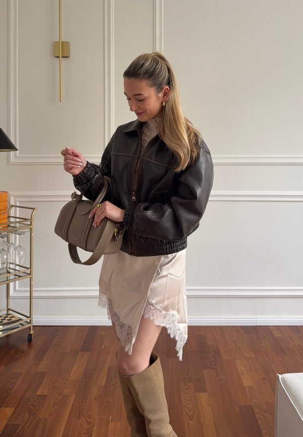 Nő hosszú hajjal, lófarokba kötve, sötét bőrdzsekit visel, bézs színű, csipkével díszített szoknyát, térdig érő csizmát hord, és taupe színű táskát tart bent.