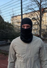 Balaclava neagră care acoperă capul și gâtul, asortată cu o jachetă bej deschis, cu fermoar, cu un finisaj texturat și un ușor luciu.