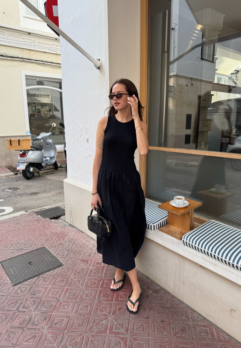 Top preto sem mangas com uma saia midi preta fluida, chinelos e uma pequena bolsa preta. Almofadas listradas num parapeito de janela de madeira.
