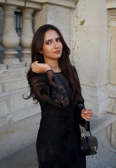 Vestido negro de manga larga con textura floral; mangas transparentes. La modelo sostiene un pequeño bolso negro con una correa de cadena plateada contra una pared de piedra.