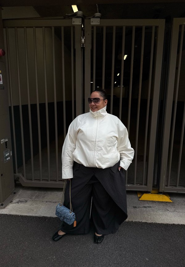 Femme portant une veste blanche et un pantalon large noir à fines rayures tient un petit sac à main bleu devant une porte métallique.