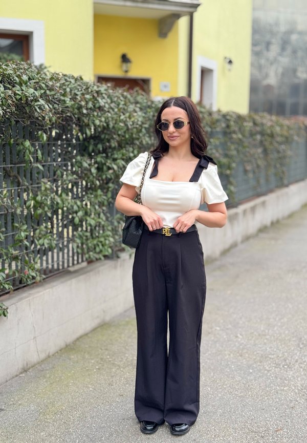 Femme portant un haut blanc à manches bouffantes et un pantalon large noir, lunettes de soleil, debout sur le trottoir près d'une haie verte et d'un bâtiment jaune.