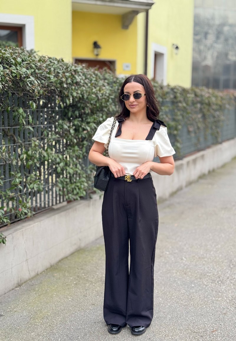 Donna che indossa una maglia bianca con maniche a sbuffo e pantaloni neri a gamba larga, occhiali da sole, in piedi sul marciapiede vicino a una siepe verde e a un edificio giallo.