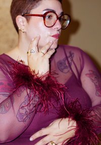 Lühikeste juuste ja prillidega inimene, kes kannab läbipaistvast lilla kangast topi sulgedega varrukatega, nähtaval on tätoveeringud ja sõrmedel kuldsõrmused.