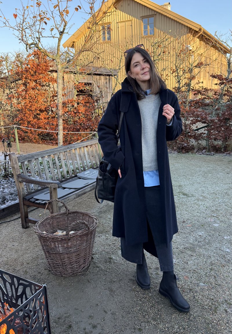 Manteau en laine noire porté sur un pull gris et une chemise bleue, associé à un pantalon sombre et des bottines noires. En arrière-plan, un banc en bois et du feuillage.