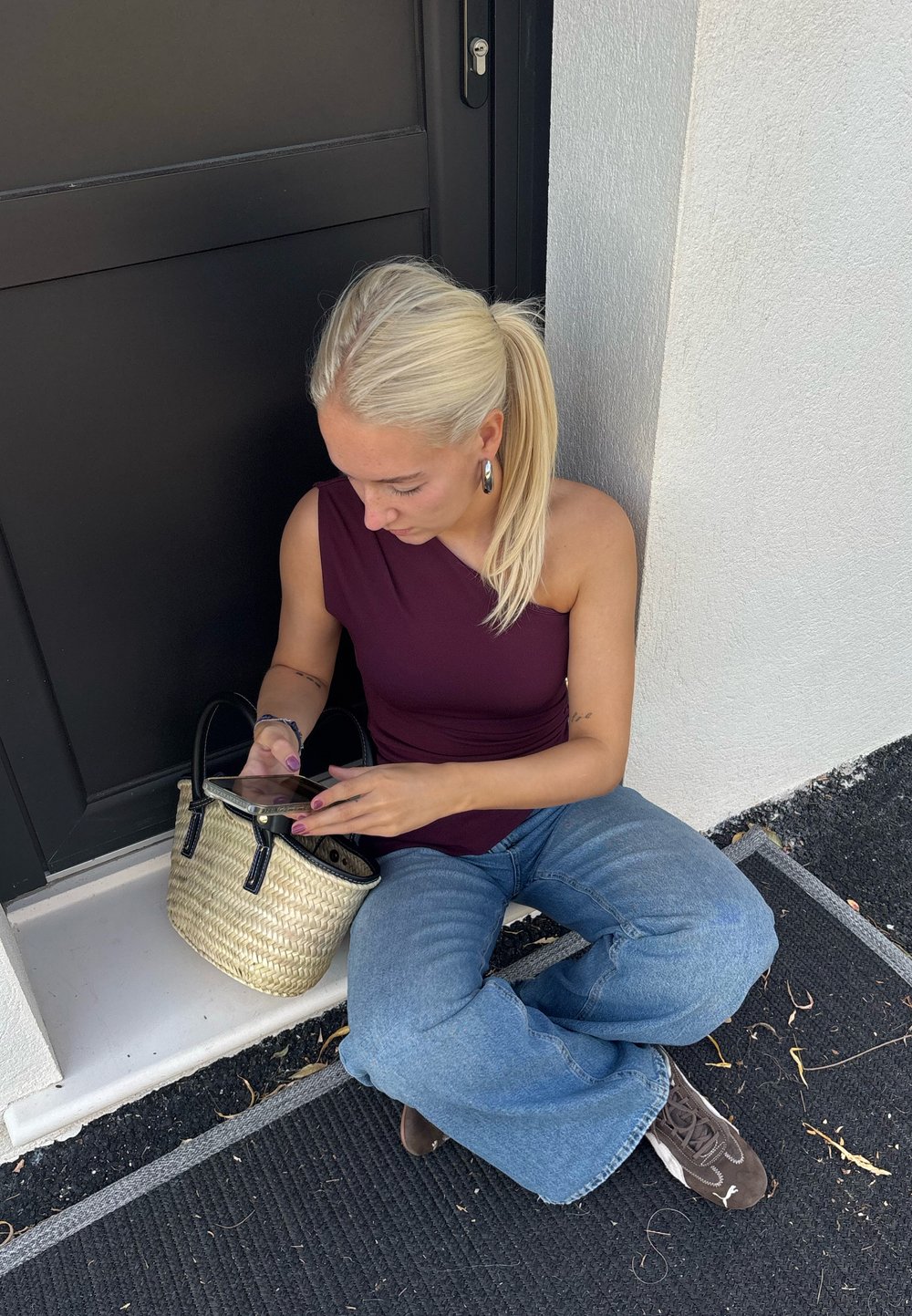 Woven straw handbag with black leather handles; osoba w bordowej bluzce na jedno ramię i jasnoniebieskich szerokich spodniach, siedząca.