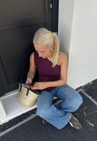 Woven straw handbag with black leather handles; osoba w bordowej bluzce na jedno ramię i jasnoniebieskich szerokich spodniach, siedząca.