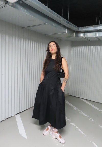 Una mujer está de pie en un aparcamiento cubierto, lleva un vestido negro sin mangas, zapatillas blancas y un pequeño bolso de mano negro.