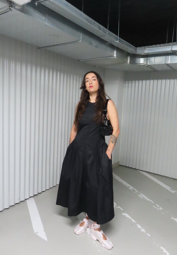 Woman stands in a parking garage wearing a sleeveless black dress, white sneakers, and carrying a small black handbag.