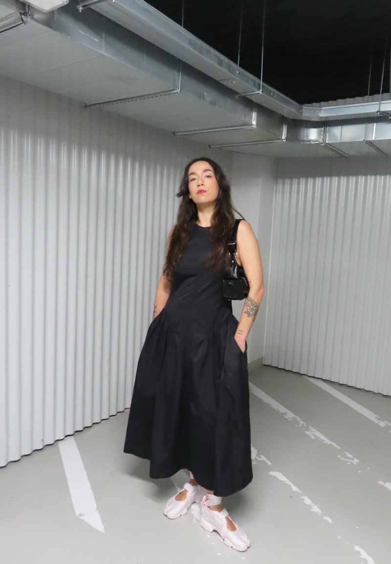 Une femme se tient dans un parking, vêtue d'une robe noire sans manches, de baskets blanches et porte un petit sac à main noir.