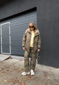 Femme portant des lunettes de soleil, veste beige doublée de fourrure, pull jaune, pantalon à imprimé léopard, baskets blanches, tenant un sac à main vert, debout près d'une porte de garage grise.