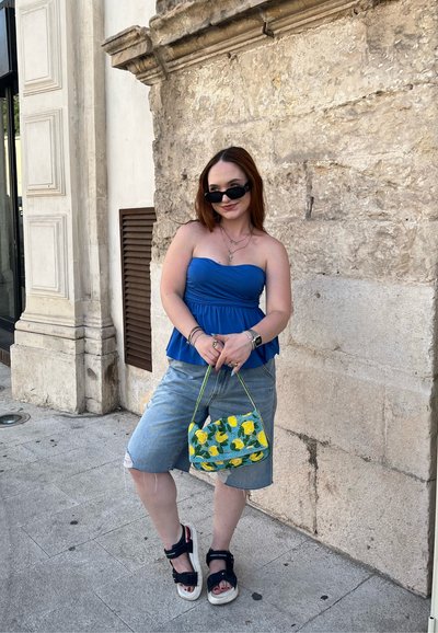 Top azul sin tirantes, shorts de mezclilla rasgados y sandalias negras. Lleva una pequeña bolsa verde con patrones de limones amarillos. La luz del sol se refleja en las superficies.