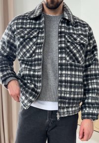 Casaco em padrão xadrez preto e branco, feito de tecido macio. Apresenta dois bolsos frontais, gola e fecho de correr na frente. Vestido por cima de um suéter cinzento.