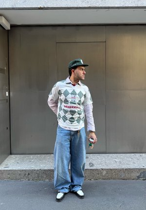 Camisa blanca de manga corta con patrón de diamantes verdes, pantalones vaqueros holgados de color azul claro y zapatos en blanco y negro. Lleva una gorra verde y sostiene una bebida.