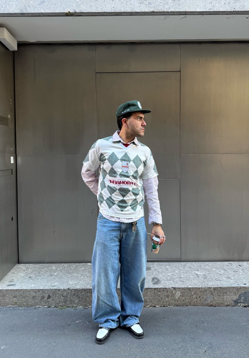 Chemise blanche à manches courtes avec un motif de losanges verts, jean baggy bleu clair, et des chaussures noir et blanc. Casquette verte et tient une boisson.
