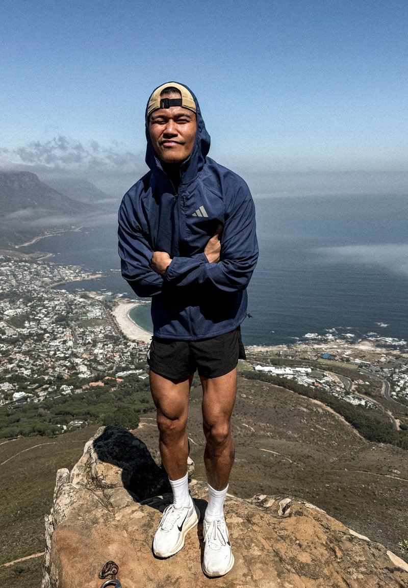 Homme en sweat à capuche bleu marine, short noir et baskets blanches, debout sur une falaise rocheuse surplombant une ville côtière et l'océan sous un ciel dégagé.
