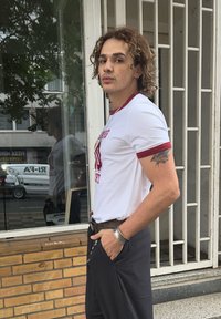 Young person with curly hair, wearing a white and red t-shirt and dark trousers, standing with hands in pockets outside a building with a window and metal bars.