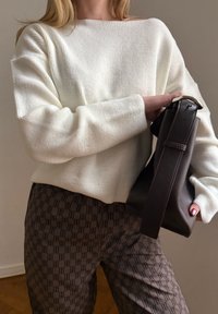 Personne portant un pull blanc en maille et un pantalon marron à motifs, tenant un sac à main marron foncé avec une boucle dorée.