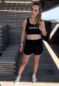 Sujeción deportiva negra con logotipo blanco, pantalones cortos negros y zapatillas de correr grises con acentos amarillos, de pie contra un fondo de madera.
