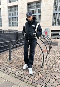 Čierno-biela varsity bunda s grafikou "TOMMY", čierne džínsy a biele tenisky. Na pozadí textúrovaný kockovaný chodník.