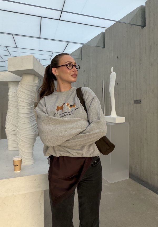 Grauer Pullover mit Hundegrafiken, lockere Passform, getragen über dunklen Jeans. Brauner Stoff über der Hüfte drapiert. Im Hintergrund sind skulpturale Elemente zu sehen.