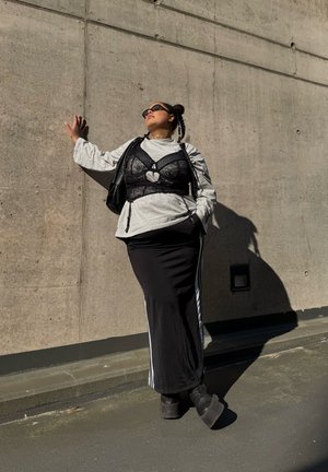 Personne avec des cheveux tressés et des lunettes de soleil s'adosse à un mur en béton, portant un haut gris, un corset en dentelle noir, une jupe noire et des bottes à plateforme.