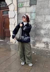 Pull polaire à motif noir et blanc, pantalon cargo olive ample, sac à bandoulière gris avec œillets, t-shirt blanc et baskets. Fond de mur en pierre.