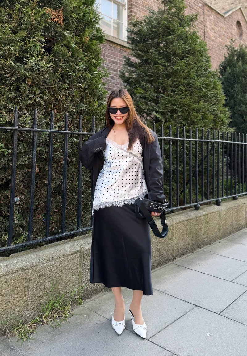 Femme portant des lunettes de soleil noires, une veste noire, un haut blanc à pois, une jupe noire et des chaussures blanches pointues, debout près d'une clôture en fer et de la verdure.