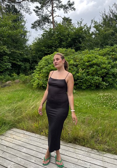 Vestido midi negro con lunares, de tirantes finos y diseño ajustado, combinado con chanclas verdes, todo ello frente a un fondo verde exuberante.