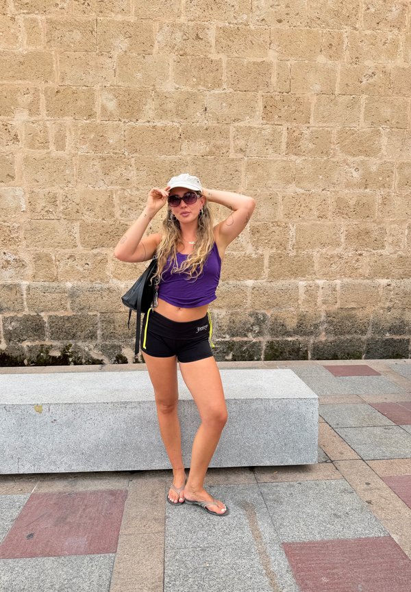 Jeune femme portant un débardeur violet, un short noir, une casquette blanche, des lunettes de soleil et des tongs, se tient sur un sol carrelé à côté d'un banc en pierre et d'un mur.