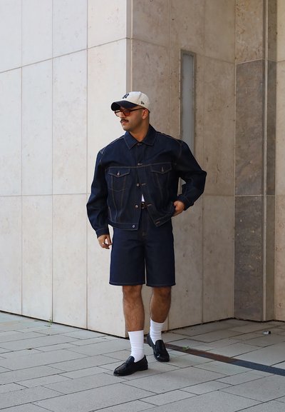 Chaqueta de mezclilla con dos bolsillos frontales, combinada con pantalones cortos de mezclilla. Zapatos negros y calcetines blancos completan el conjunto. Fondo neutro.