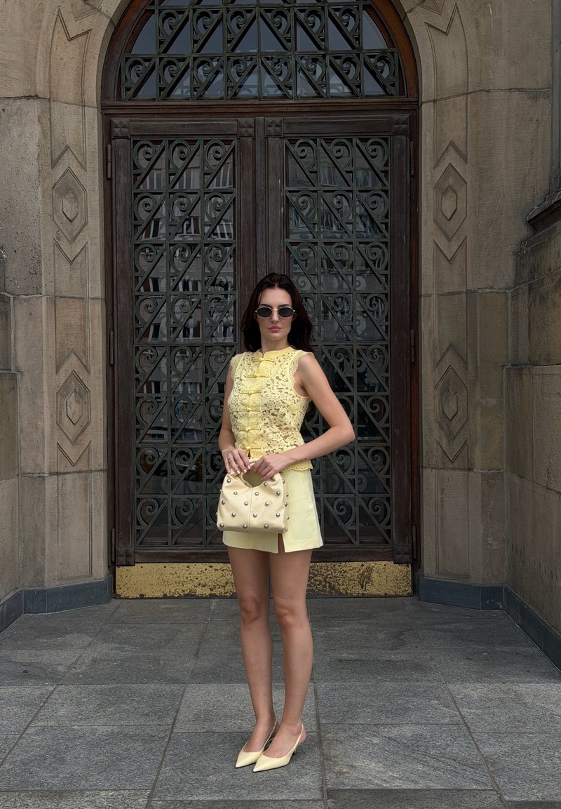 Donna con top in pizzo giallo e gonna, tiene in mano una borsa borchiata, indossa occhiali da sole e tacchi, in piedi davanti a un cancello in metallo scuro decorato.