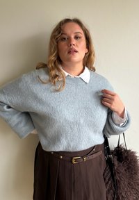 Hele sinine kootud kampsun röstitud mustriga, kombineeritud valge kraega särgi ja pruuni volditud seelikuga, millel on kuldse pandlaga vöö.