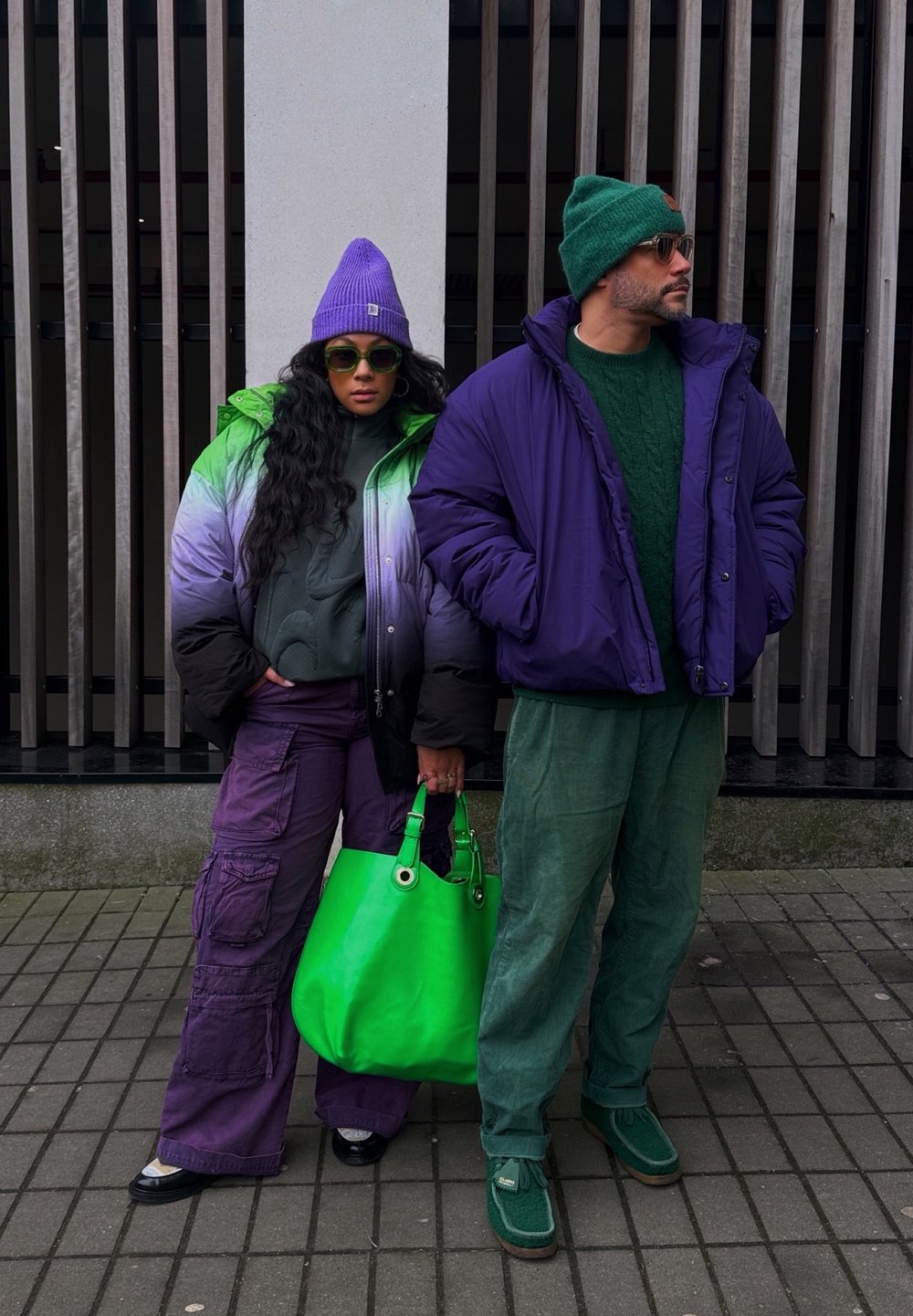 Δύο άτομα στέκονται δίπλα-δίπλα. Η γυναίκα φοράει ένα μοντέρνο μπουφάν με πράσινο και μοβ gradation, μωβ cargo παντελόνι και κρατά μια φωτεινή πράσινη τσάντα. Ο άνδρας φοράει ένα μπουφάν σε μοβ απόχρωση και πράσινο πλεκτό σκουφάκι, σε συνδυασμό με πράσινο πουλόβερ και παντελόνι.