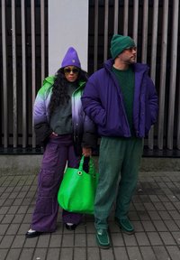 Δύο άτομα στέκονται δίπλα-δίπλα. Η γυναίκα φοράει ένα μοντέρνο μπουφάν με πράσινο και μοβ gradation, μωβ cargo παντελόνι και κρατά μια φωτεινή πράσινη τσάντα. Ο άνδρας φοράει ένα μπουφάν σε μοβ απόχρωση και πράσινο πλεκτό σκουφάκι, σε συνδυασμό με πράσινο πουλόβερ και παντελόνι.