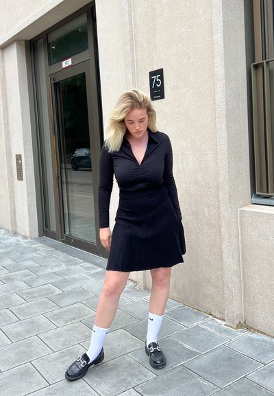 Blusa negra de manga larga con cuello y detalles fruncidos, combinada con una falda negra de canalé. Mocasines negros y calcetines blancos.