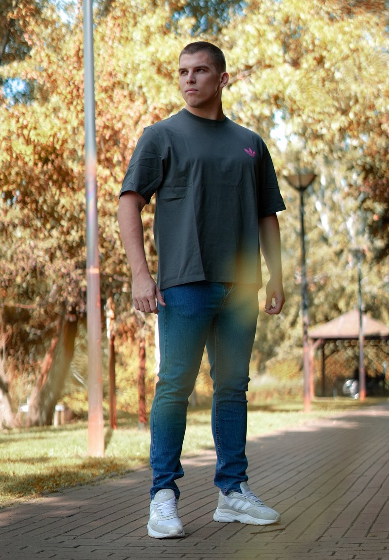Grå bomullströja med korta ärmar och rosa logga, tillsammans med blå jeans och vita sneakers, stående i en park.