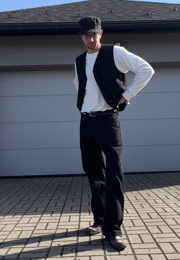Hombre con gorra negra, gafas, chaleco negro, camisa blanca de manga larga, pantalones y zapatos negros, de pie en un camino pavimentado frente a una puerta de garaje.