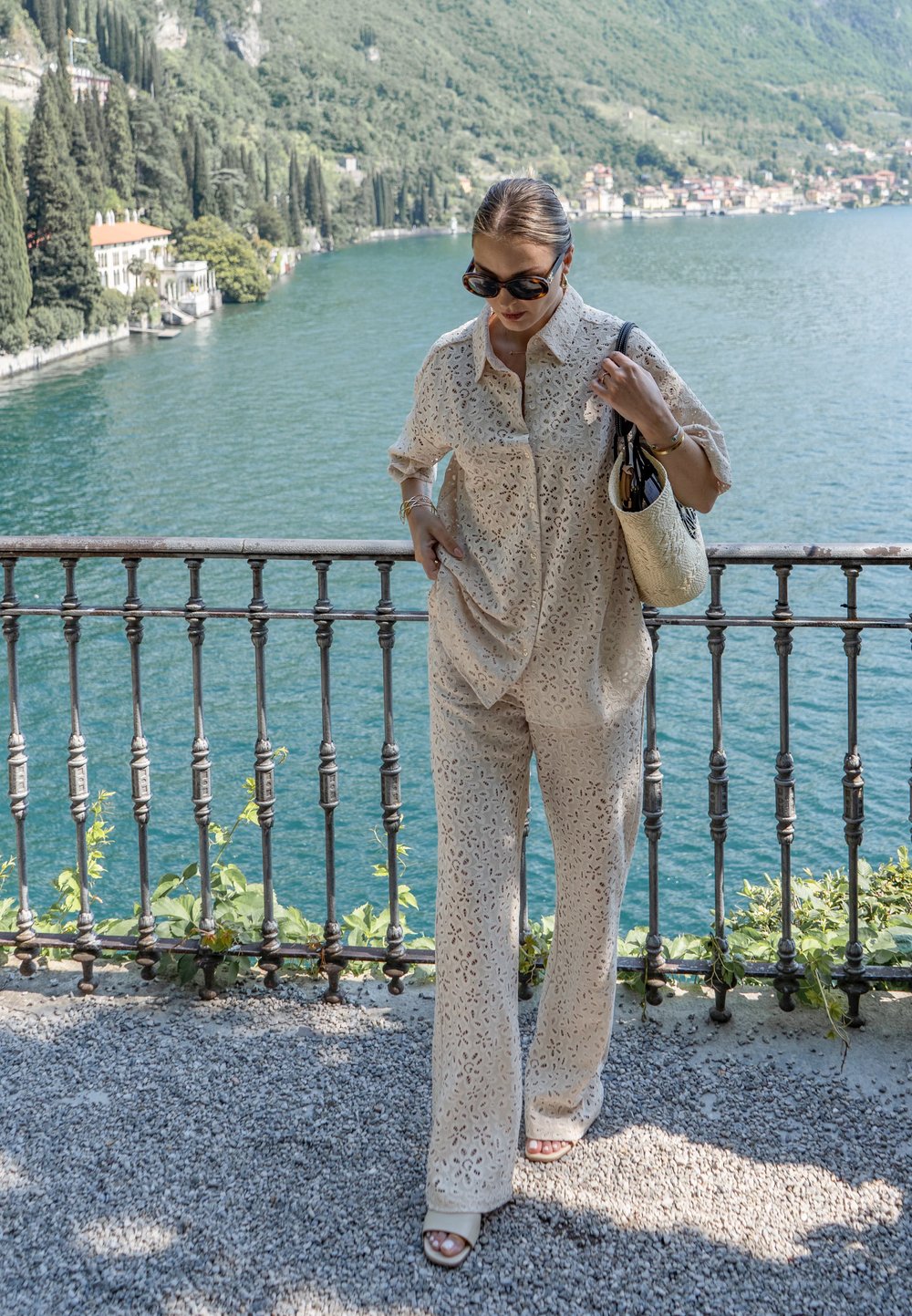 Donna che indossa un outfit in pizzo beige e occhiali da sole, con una borsa intrecciata, che cammina lungo una ringhiera sul lago con montagne e acqua sullo sfondo.