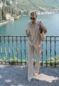 Donna che indossa un outfit in pizzo beige e occhiali da sole, con una borsa intrecciata, che cammina lungo una ringhiera sul lago con montagne e acqua sullo sfondo.