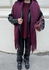 Écharpe en laine bordeaux avec franges, veste en cuir noire, pull rouge foncé et pantalon en similicuir noir. Petit sac bordeaux à la main.