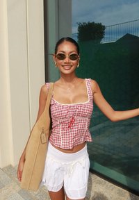 Jonge vrouw met zonnebril, rood geruit mouwloos topje, witte shorts, die een tan schoudertas draagt, staand in de buitenlucht bij een glazen raam.