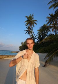 White, loose-knit shirt with a deep neckline, worn over white shorts, paired with a black watch. Tropical beach setting with palm trees.