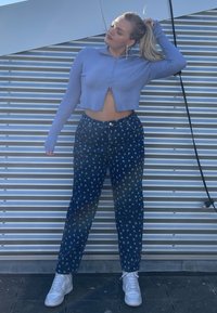 Jovem mulher de pé contra uma parede de metal ondulado, vestindo um suéter azul cropped, jeans escuros com padrão, tênis brancos, segurando o cabelo em um rabo de cavalo.