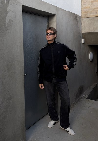 Chaqueta negra con cierre, adornada con tres rayas grises, combinada con jeans negros holgados y zapatillas blancas con acentos negros, frente a una pared gris texturizada.