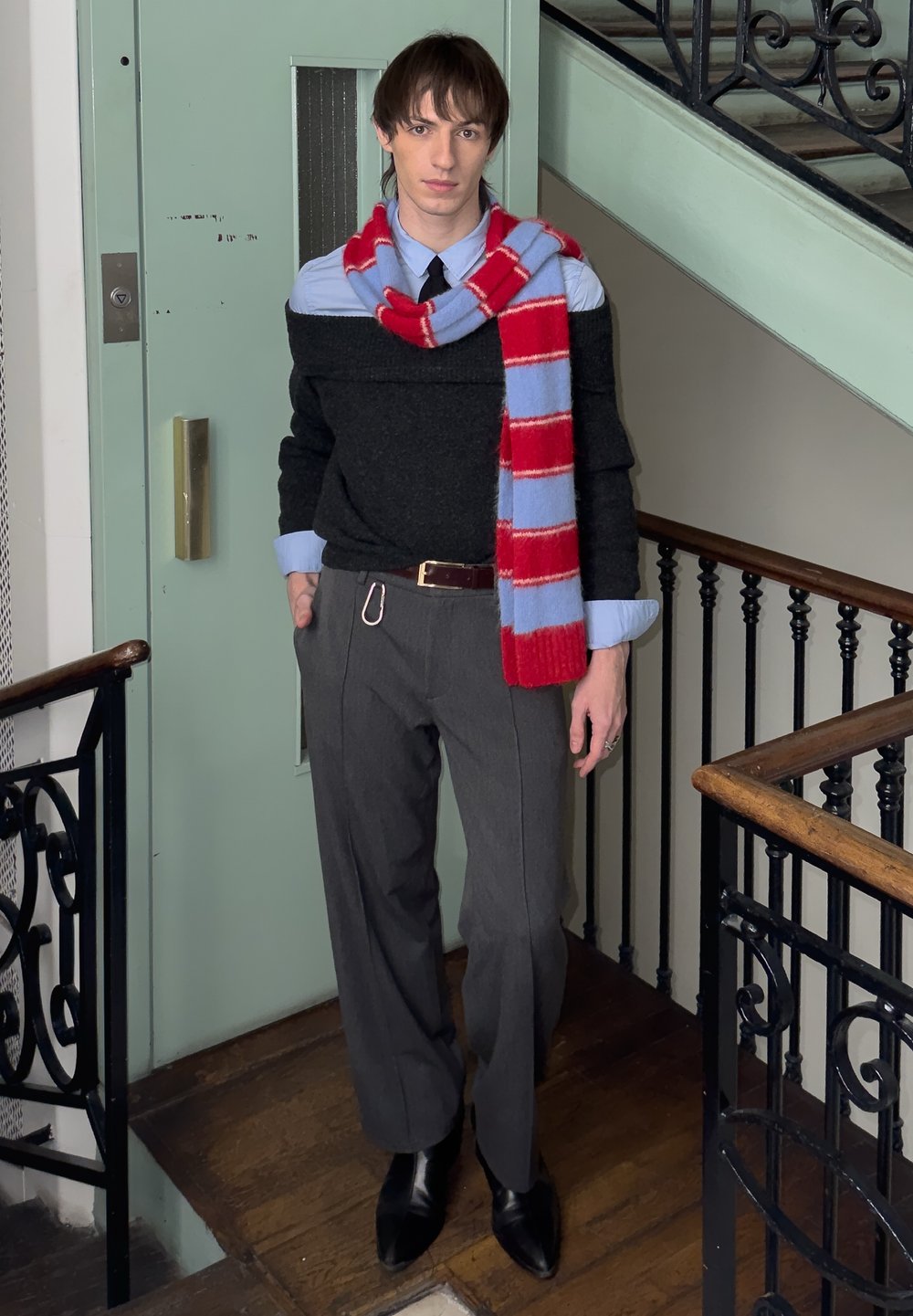 Jeune personne debout à l'intérieur près d'une rampe d'escalier, portant un pantalon gris, un pull noir à épaules dénudées par-dessus une chemise bleue, et une écharpe rouge et bleue à rayures.
